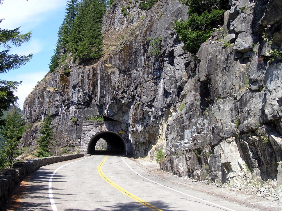 A road passes though an arched tunnel that cuts through a rocky hillside.