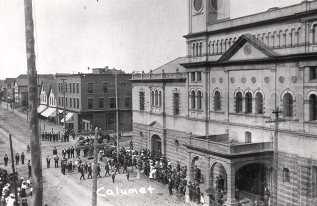 Historic photo of a crowd of people in the street and sidewalk beside a large, ornate theatre.