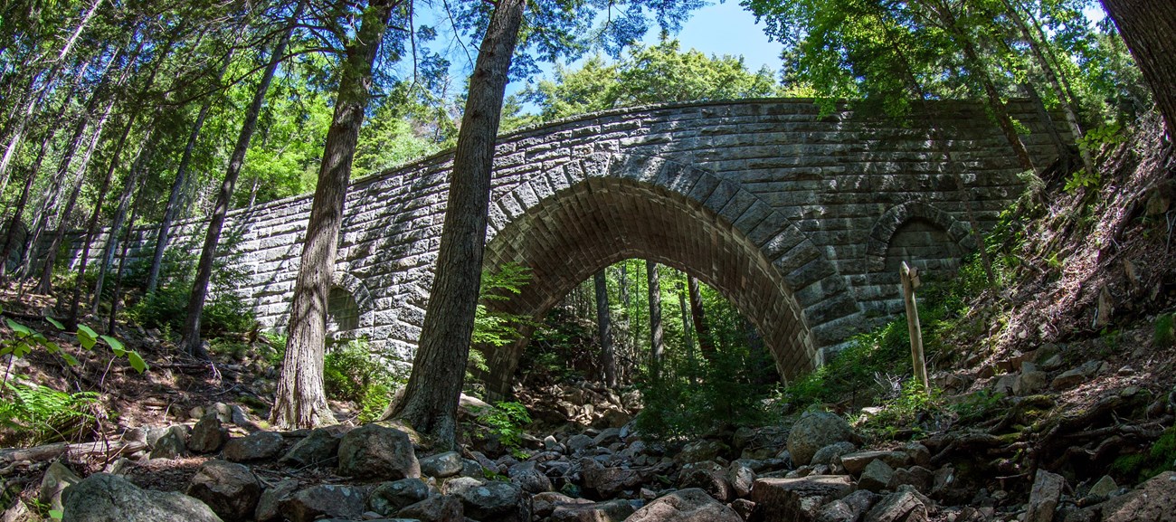 A cobblestone bridge with a high arched opening crosses over a dry, rocky creek bed in a wooded area