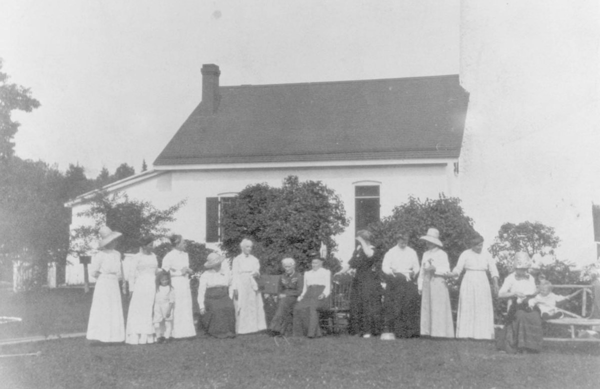 A line of women in long dresses in front of the Michigan Island Light Station, early 1900s