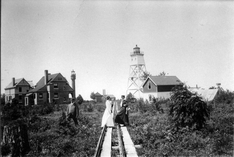 Three people stand on tram tracks beside the buildings and a lighthouse