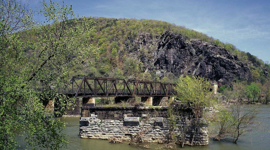 A railroad bridge with metal trusses on piers crosses a river, beside a rocky and tree-covered hill.