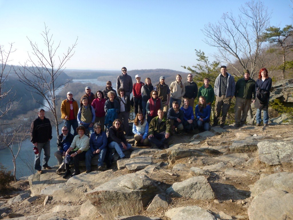 A group photograph on a rocky, sunny overlook framed by leafless trees and a view of a wide river