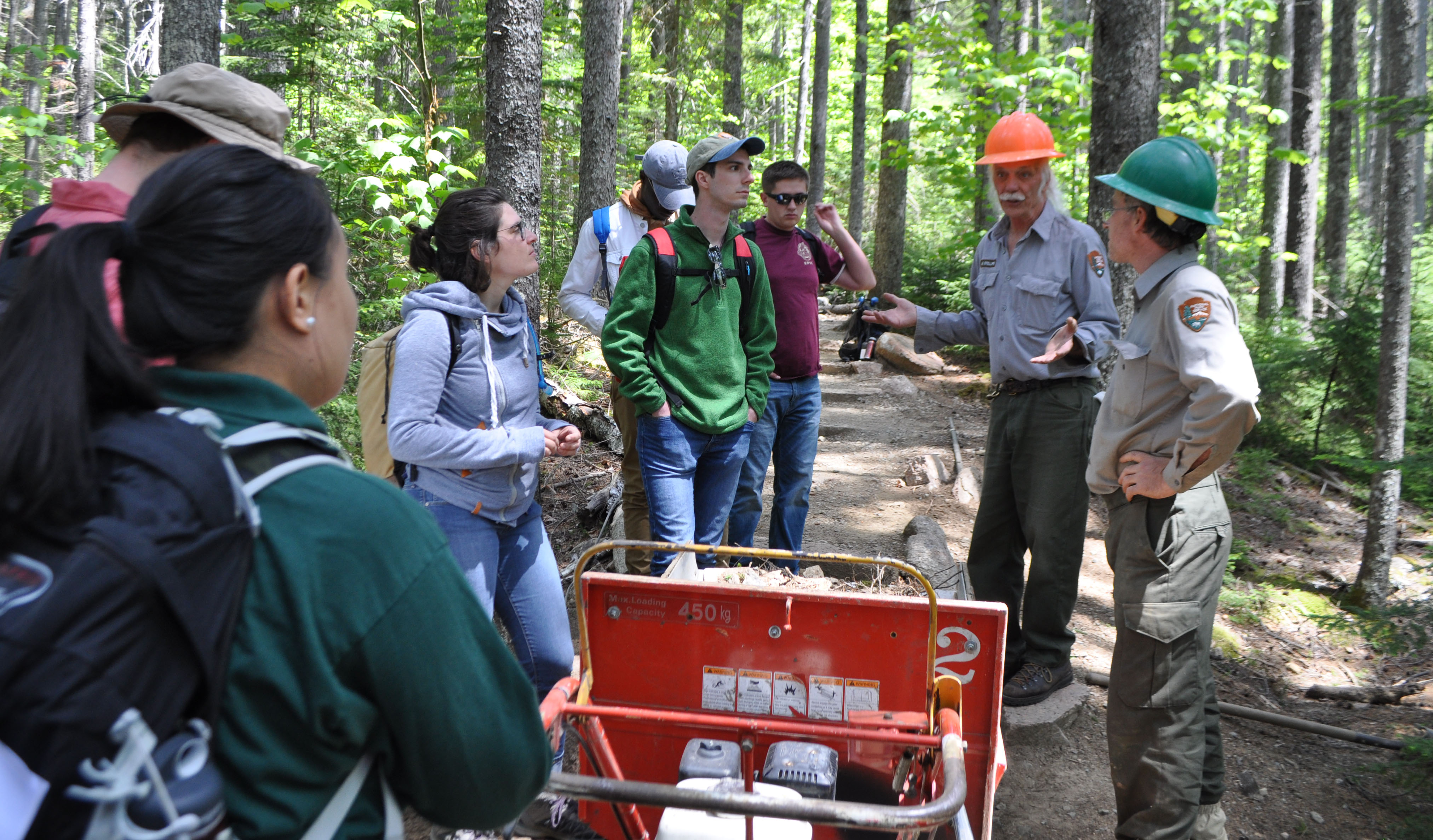 A group of young adults gathers to listen to two maintenance employees in hard hats, standing along a trail through shady woods