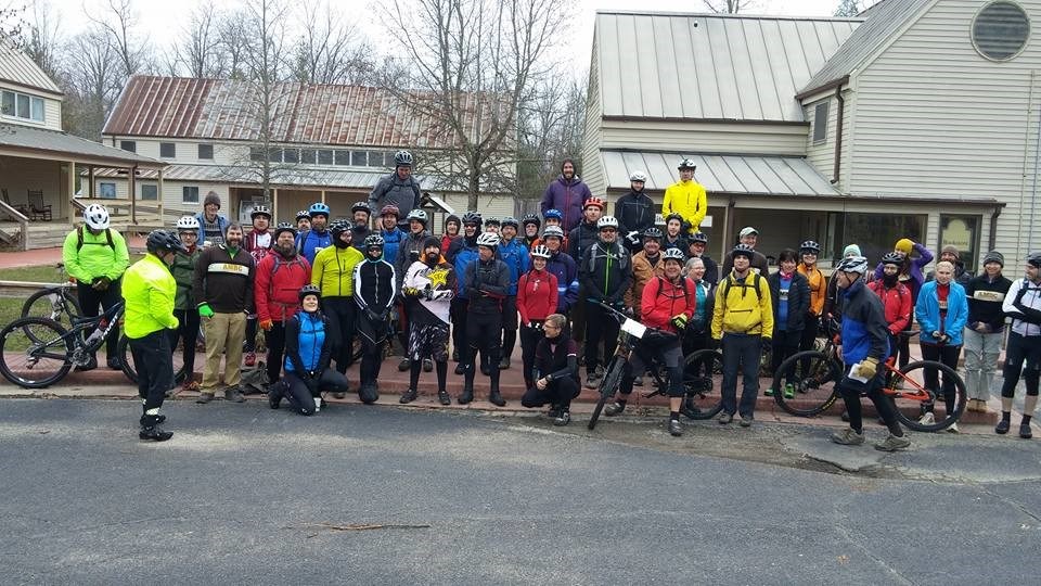 A large group of cyclists in helmets, bright clothes pose in front of several barn-like structures