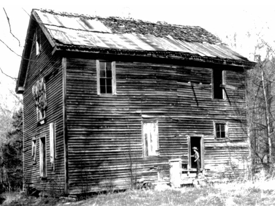 A black and white historic photo of a boy in the doorway of the Boxley Grist Mill, a two-story structure with wood siding