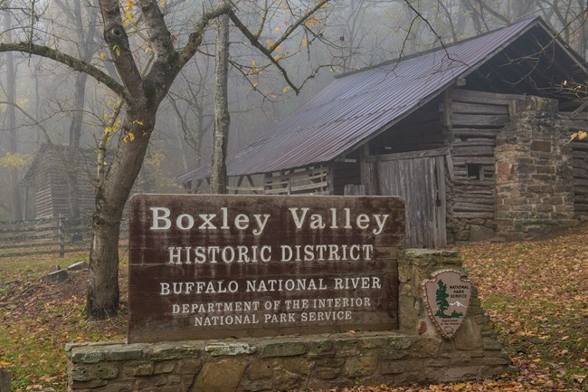 A National Park Service sign that reads, "Boxley Valley Historic District," with a historic log and stone structure in the background.