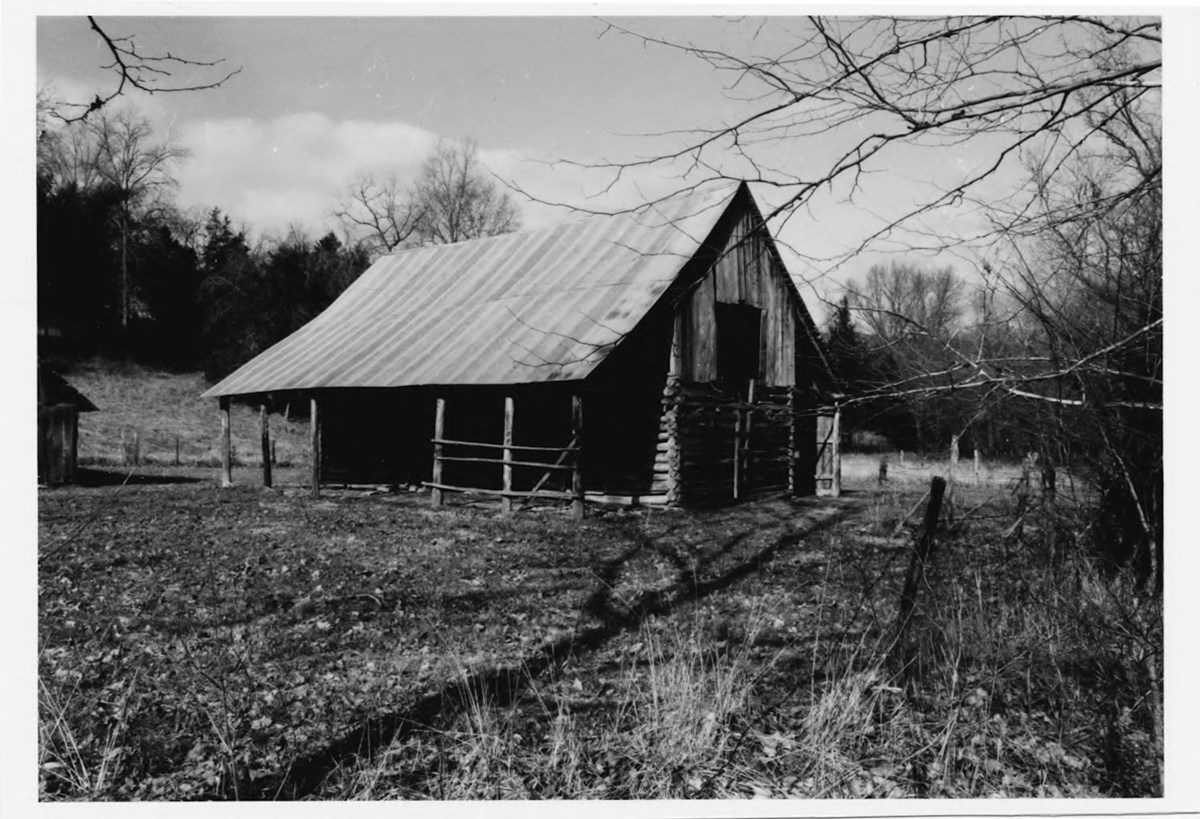 Black and white photo of log barn with a metal roof, surrounded by open yard and leafless trees