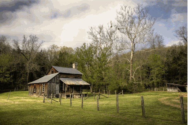 A log cabin in a rural landscape, surrounded by open turf with fencing and woods in the background