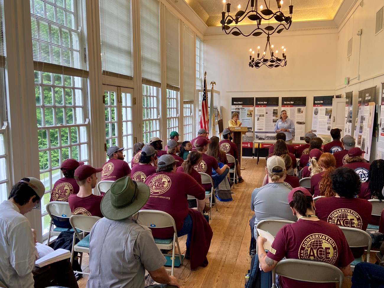 LSC Baltimore in Classroom A group of young adults are seated in folding chairs in a narrow room with tall, many-paned windows and chandeliers, facing two presenters in front.