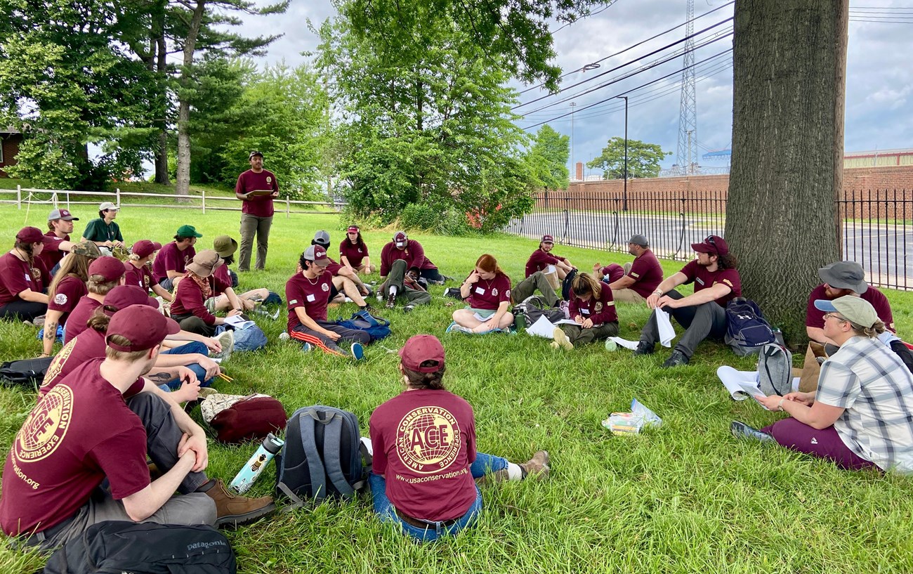 LSC Baltimore group under tree A group of young adults, most in red shirts and ball caps, are seated in grass at the base of a tall tree near a metal fence.