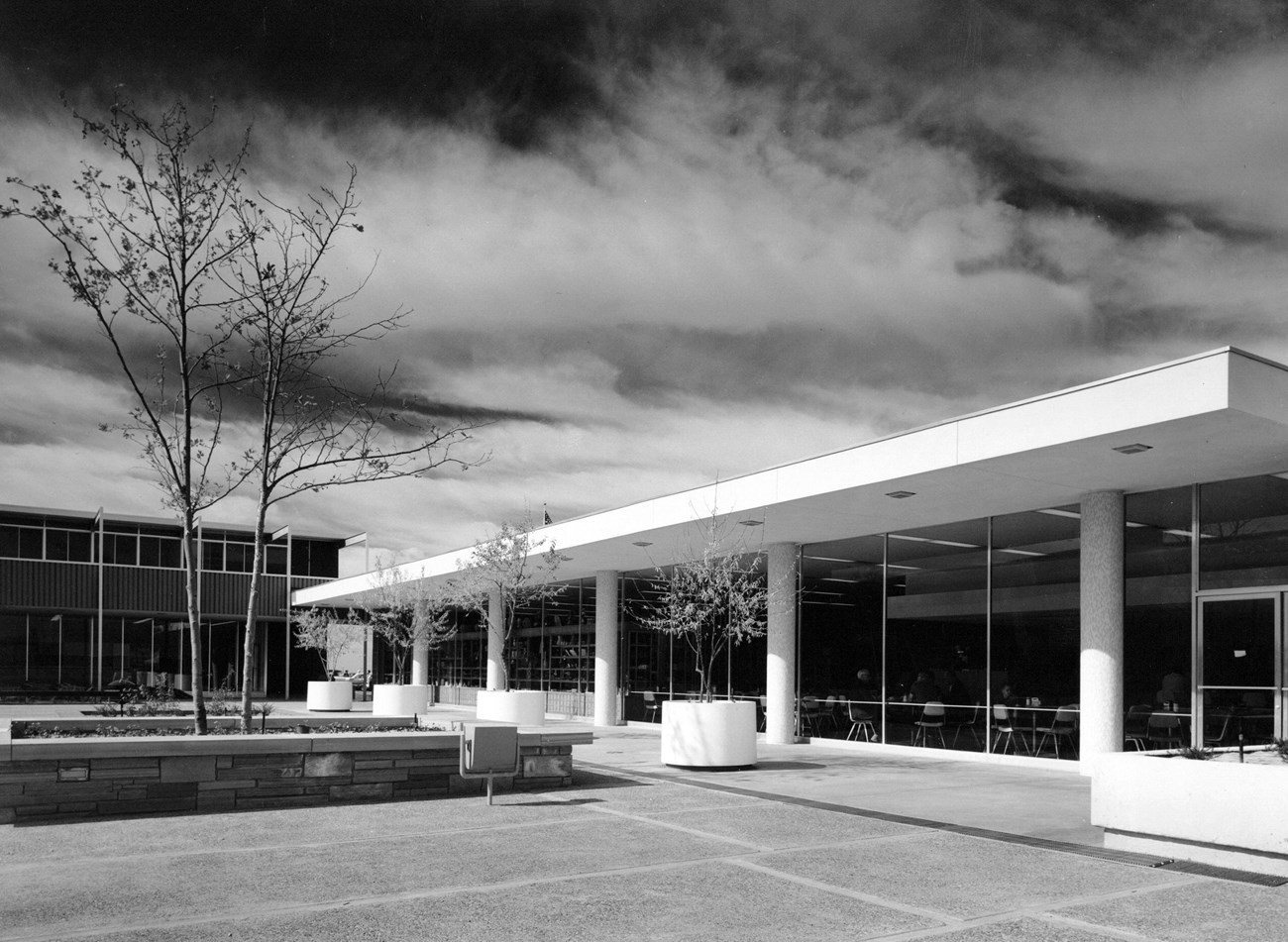 A row of columns support a roof overhang of a long, low building in the Painted Desert Community Complex, facing the open paved plaza with a square planter in the middle.