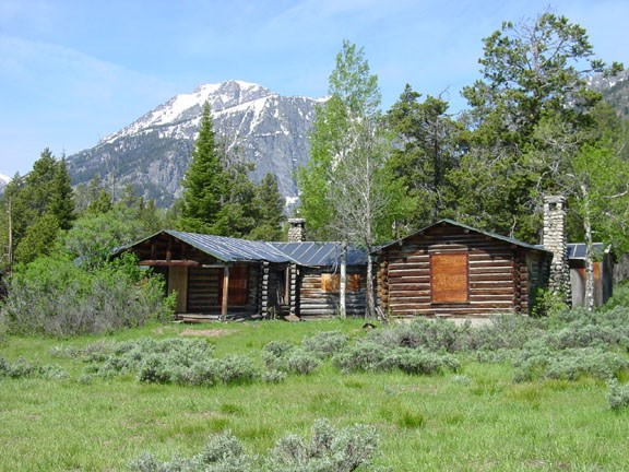 Best Hammond cabin 6-5-04 - full Single-story log cabin with boarded windows is surrounded by scrub, trees, and snow-capped mountains.