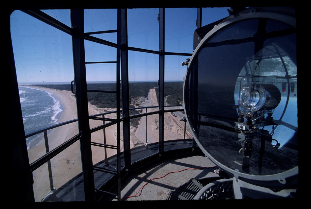 A view from behind the light at the top of lighthouse, with the sandy shoreline to the left and a clear path through trees ahead