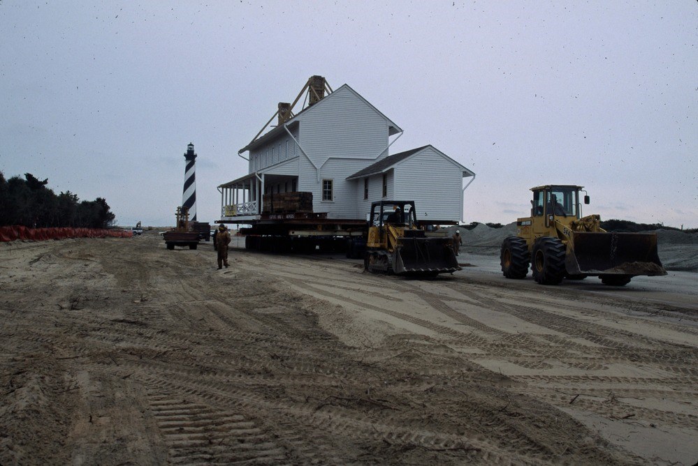 A two-story house with a porch is elevated and moved along a compacted dirt track, with bulldozers at the lead and a lighthouse in the background