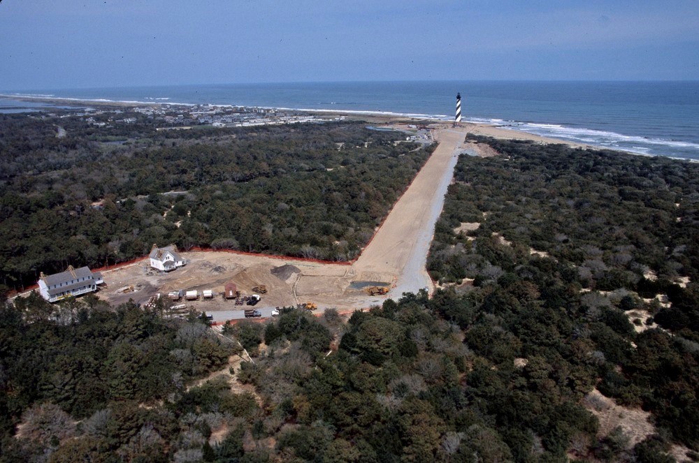 The Cape Hatteras Lighthouse stands beside the ocean and at the end of a long, straight clearing through the trees