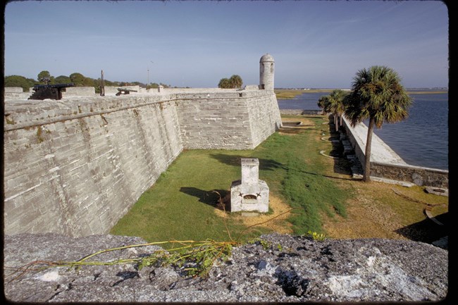 A stone furnace in an area of turf beside the stone walls of a fortification, beside water