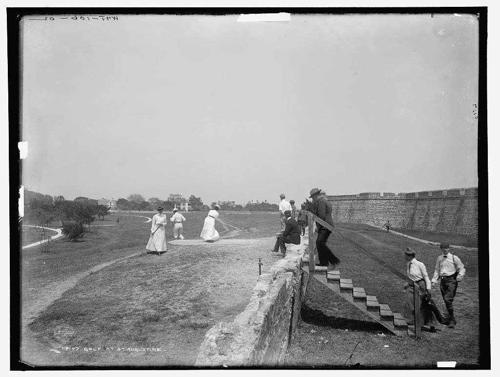 A group of golfers in 1902 on an area of turf, beside wooden stairs over a stone defensive structure. A fort is in the background right
