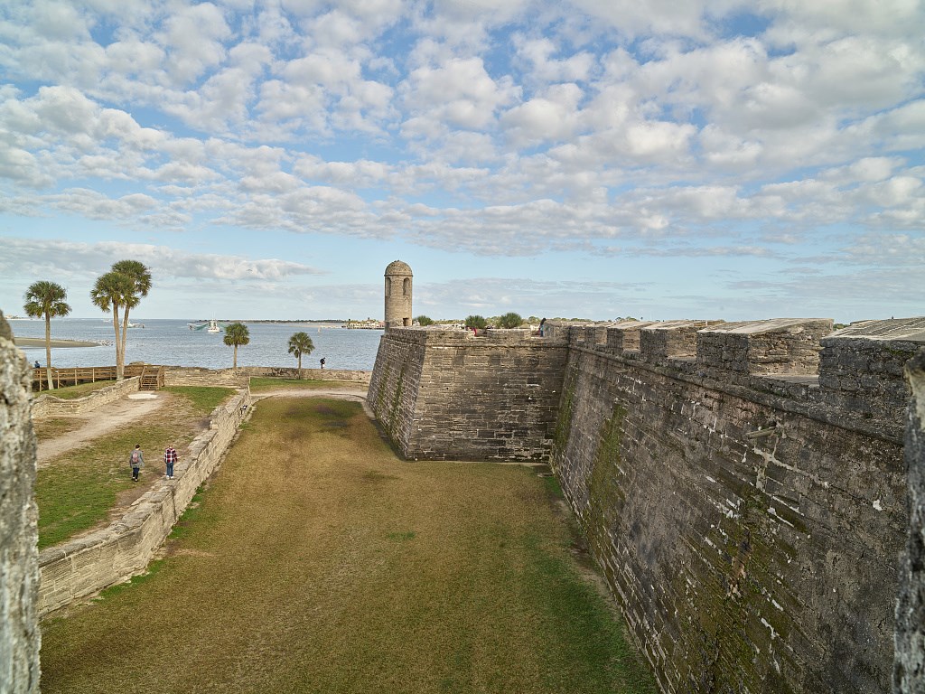 A stone wall of a fortification rises beside an area of turf and defensive works, with a bay in the background