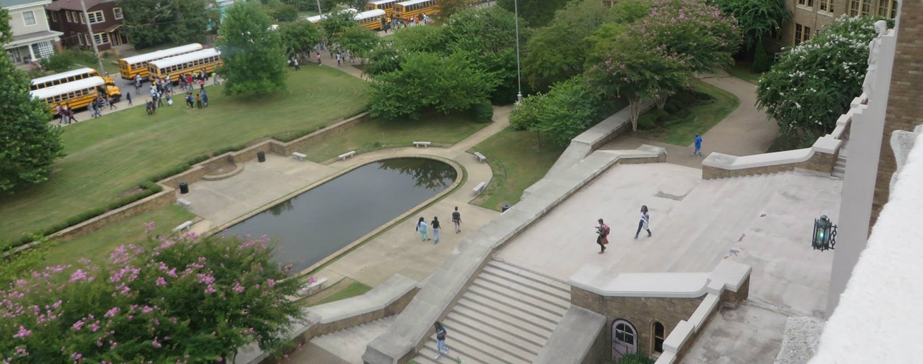 Little Rock Central High School NHS Landsape in front of a brick school includes an oval reflecting pool, trees and turf, entrance staircase, buses, and row of houses across the street