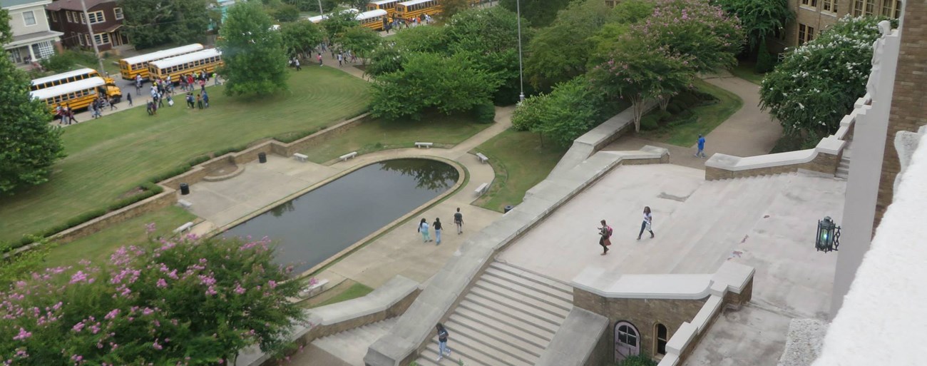 Landsape in front of a brick school includes an oval reflecting pool, trees and turf, entrance staircase, buses, and row of houses across the street