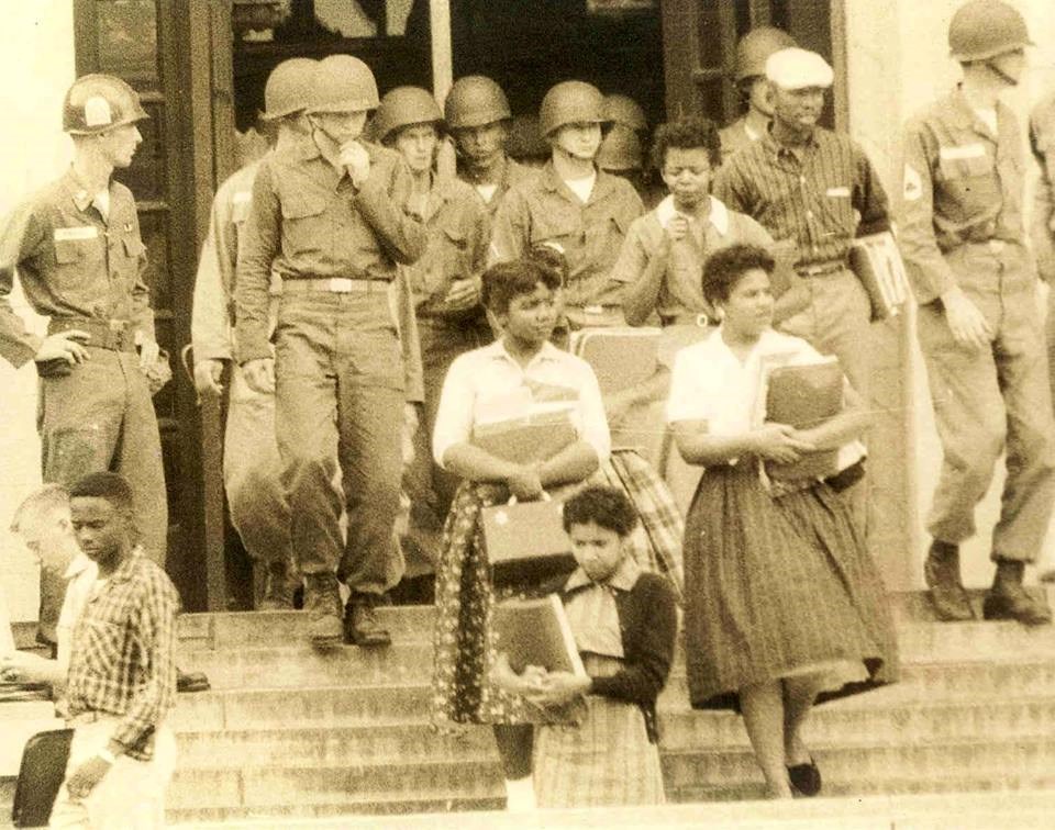 Historic photograph of four African American young women holding school books, walking down the steps of a school building, surrounded by guards in uniform and helmets
