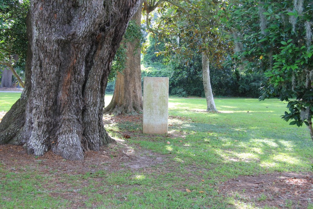 A rectangular memorial, light colored with green lichen, stands in grass under towering trees.