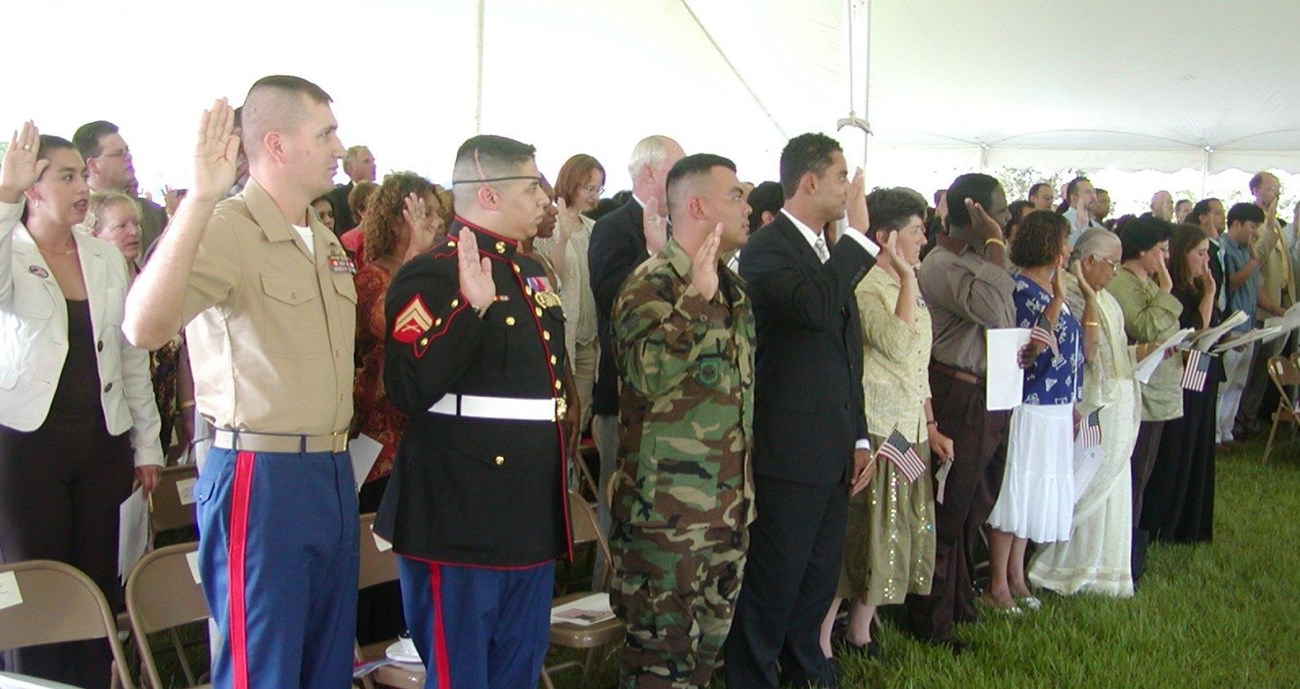 Rows of people stand beneath a tent, with right hands raised