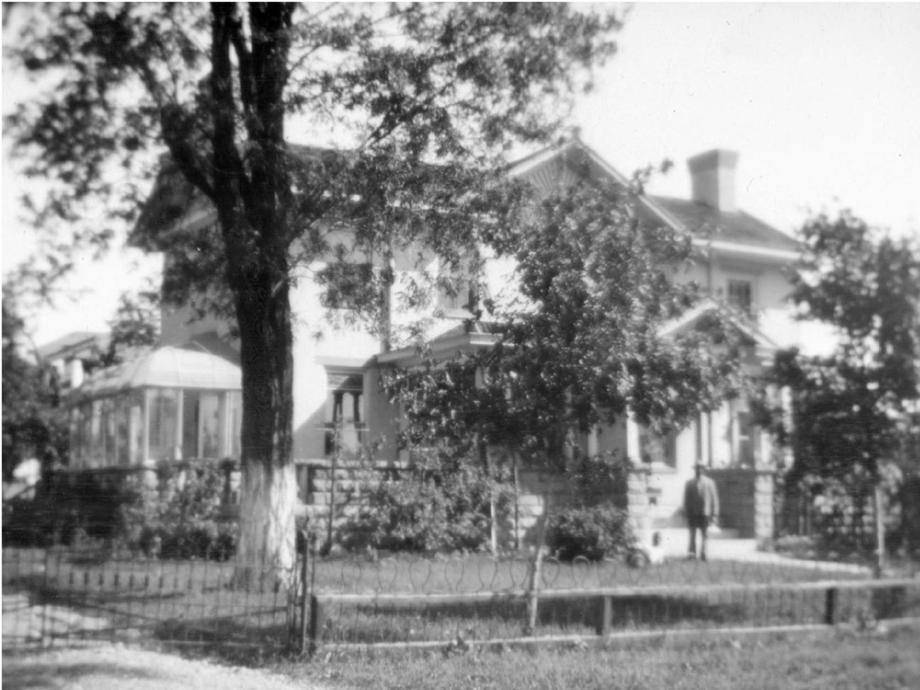 CHYO 1910 front of house A man stands near the front porch of a two-story house, in a fence-enclosed yard with trees, garden beds,