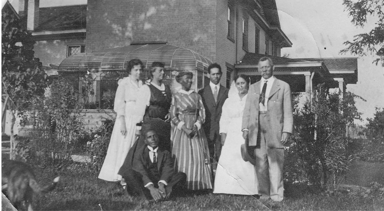 CHYO group posing in garden A group of seven people, in dresses and suits, pose beside a row of shrubs. A house with greenhouse is in the background.