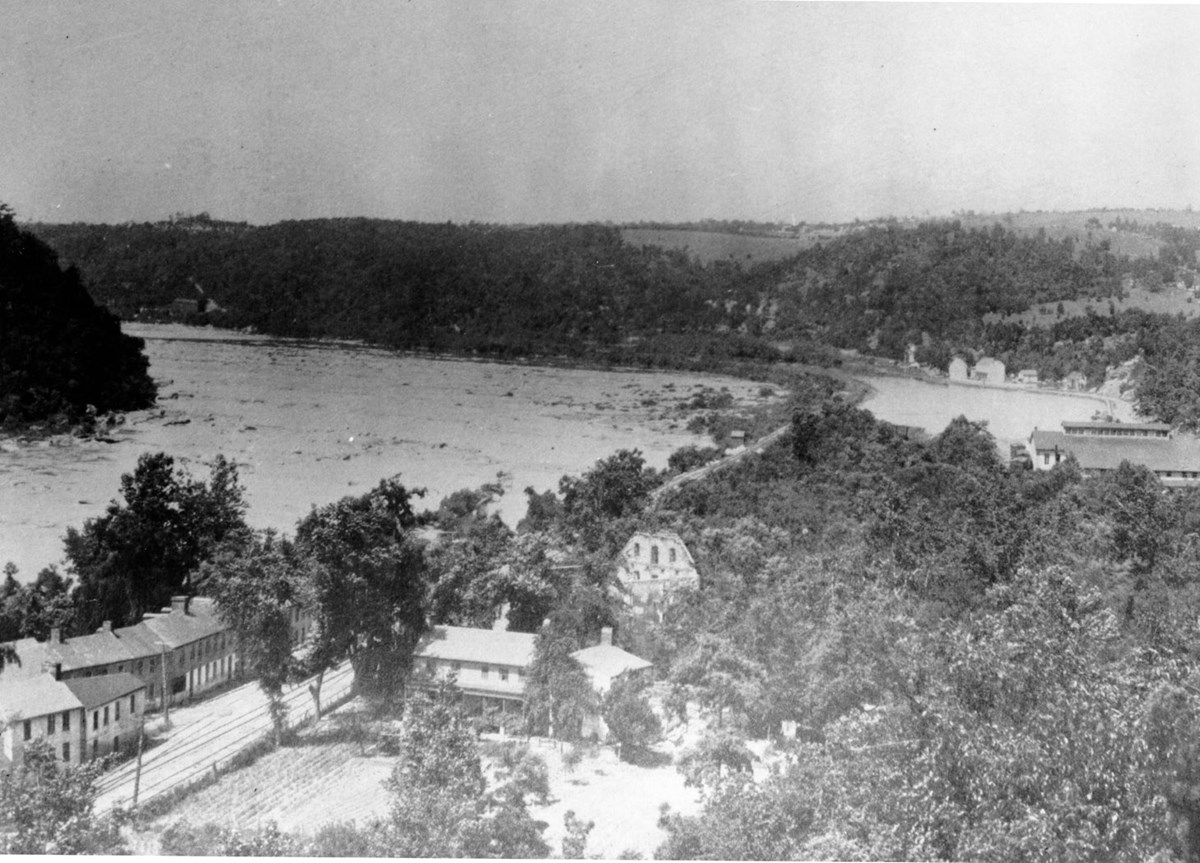 Black and white image from a high angle shows structures, a road or railroad, and trees on an island in a river.