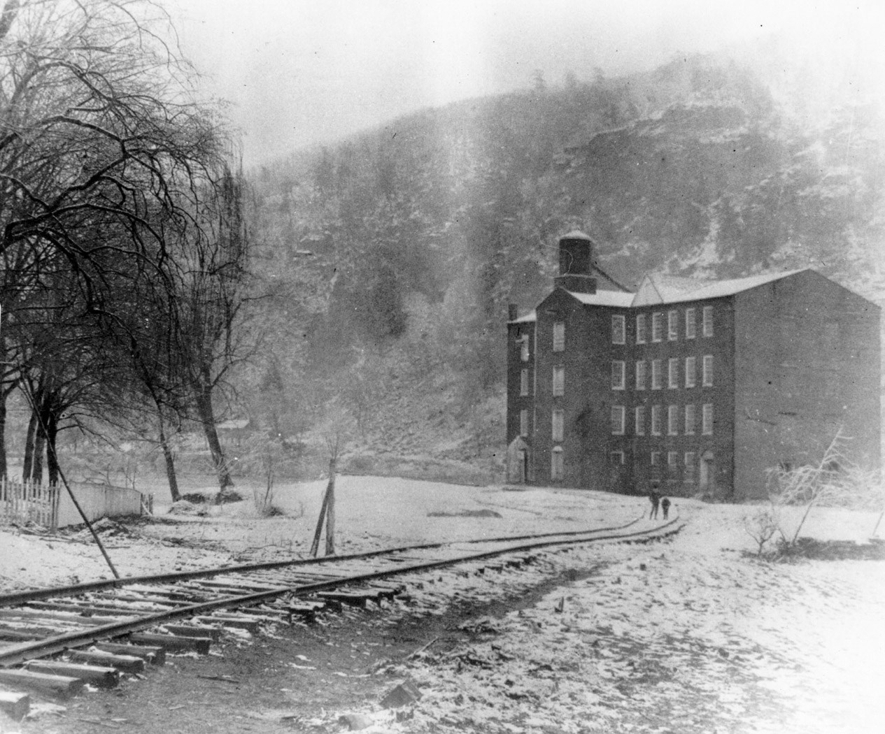 Black and white photo of a four-story mill with rows of windows stands at the end of a railroad spur, with a hillside in the background.