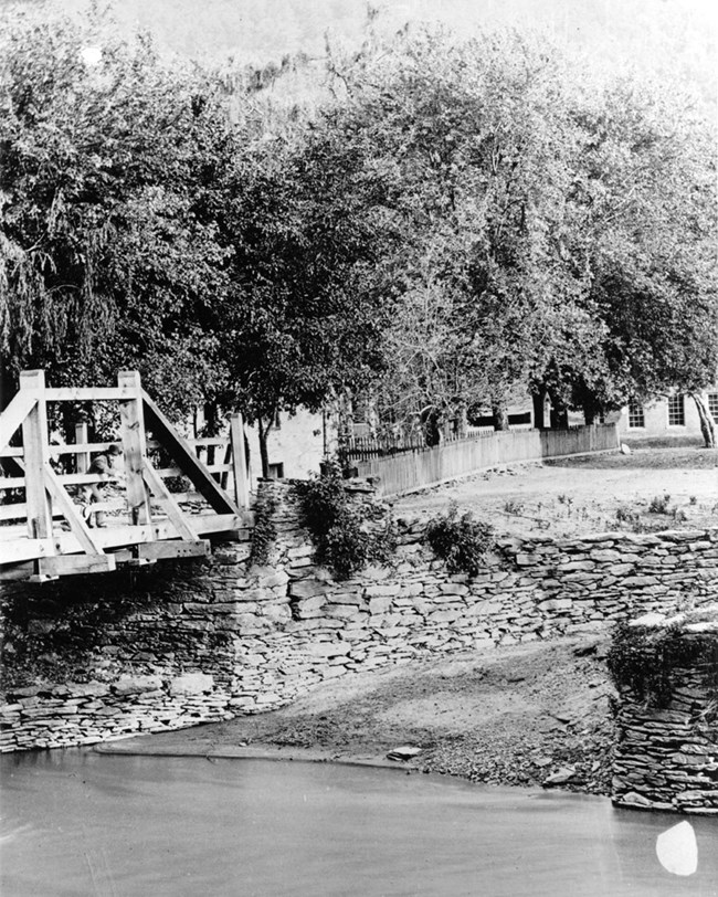 A wooden bridge crosses water to a stone retaining wall, with houses and trees on the land beyond.