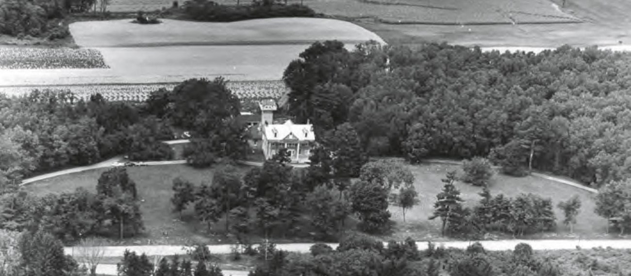 An aerial perspective of a mansion on a curved driveway in an agricultural setting. A dense woodlot, the former site of the orchard, is to the rear right.