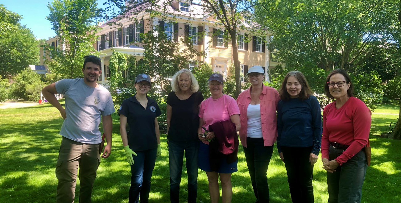 Seven people, staff and volunteers, stand in a shady lawn and garden area beside a two-story yellow house.