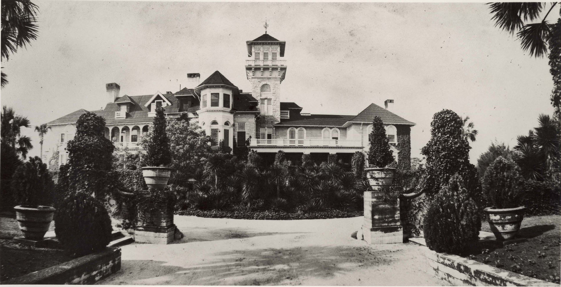 Historic image: Driveway to a large estate home is framed by landscaping, including pruned shrubs in planters.