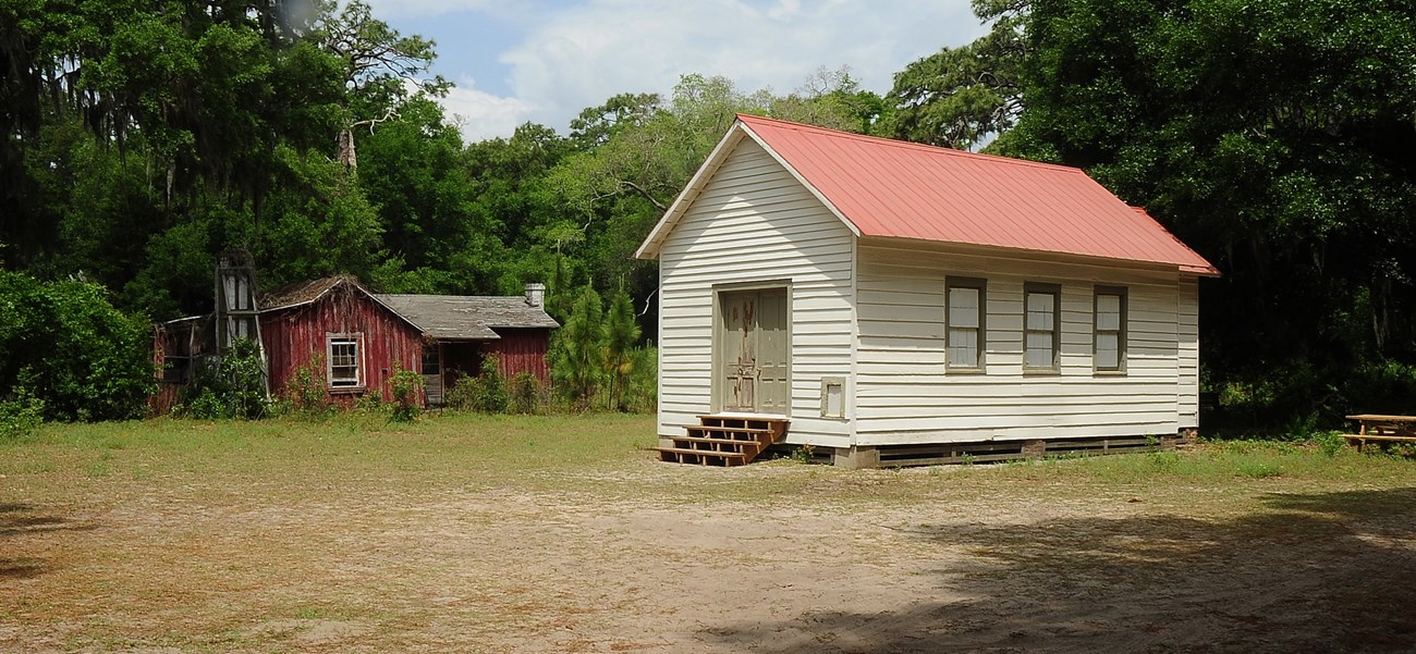 Small rectangular building with double doors at one end and white siding, with one-story dwelling surrounded by vegetation in background