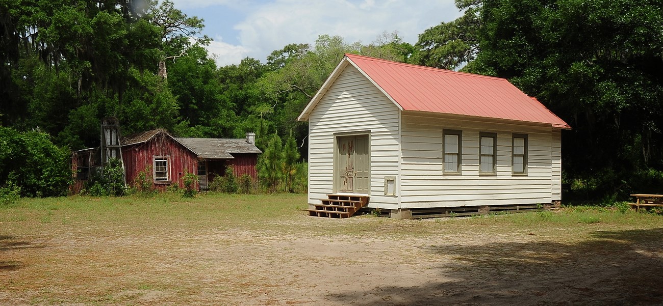 CUIS_NorthEnd-Baptist Small rectangular building with double doors at one end and white siding, with one-story dwelling surrounded by vegetation in background