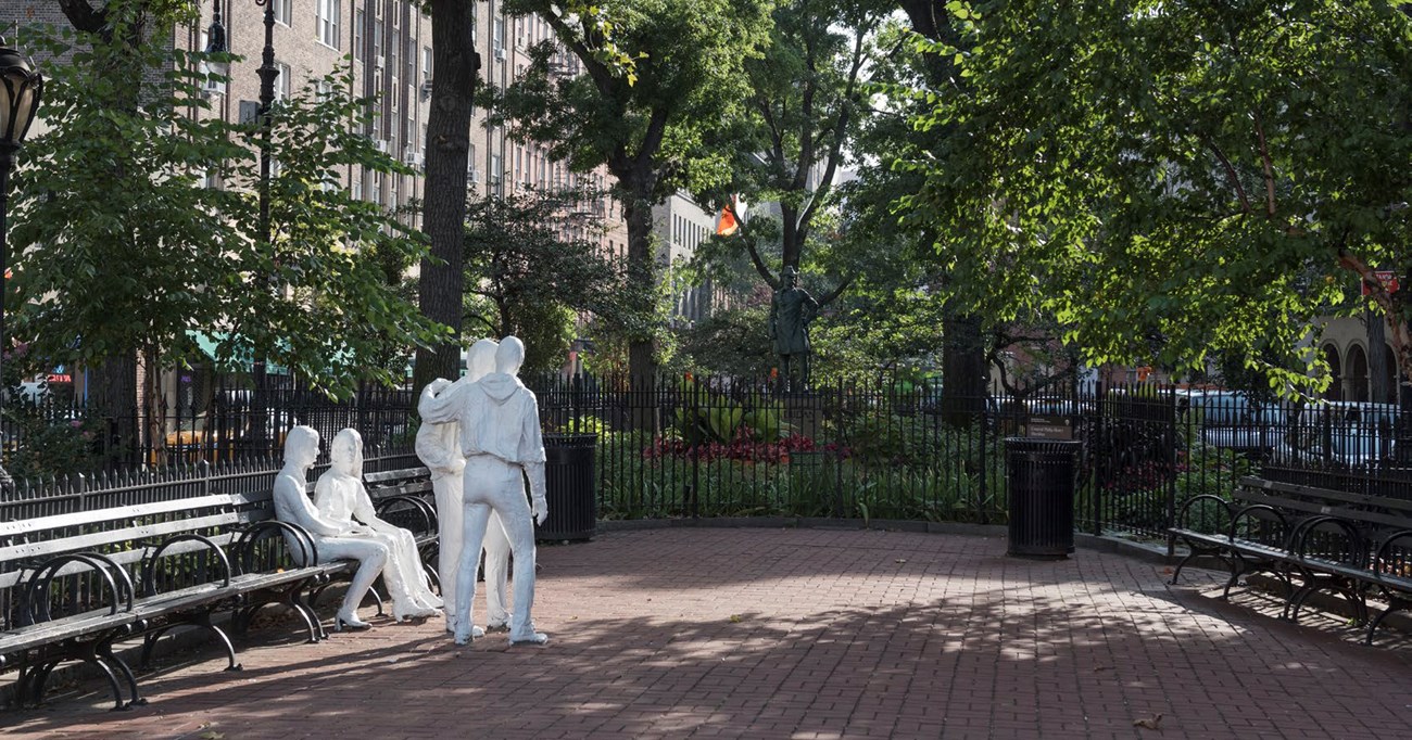 A city park with trees, benches, and fencing also contains a sculptures of four people, two seated and two standing by the bench.