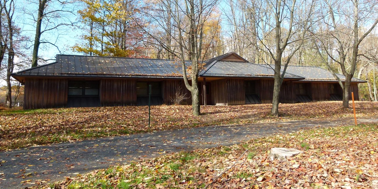 A driveway and line of trees in front of a long, one-story modern camp dorm in fall.