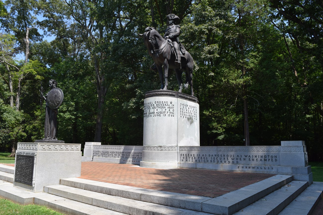 The Nathanael Greene Statue is a man on a horse atop a monument base with writing, with a brick viewing platform and smaller monument in front.