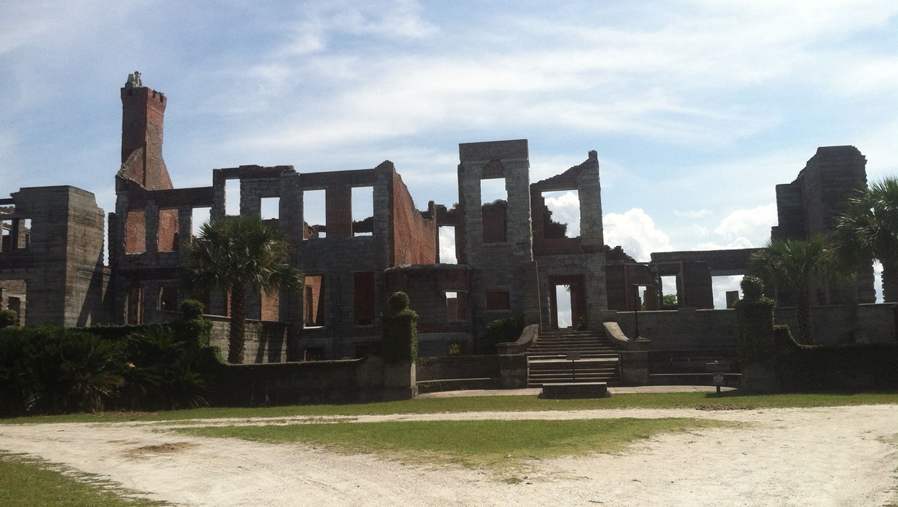 Vines cover sections of a two-story building ruin with a brick chimney and rectangular holes that were once windows.