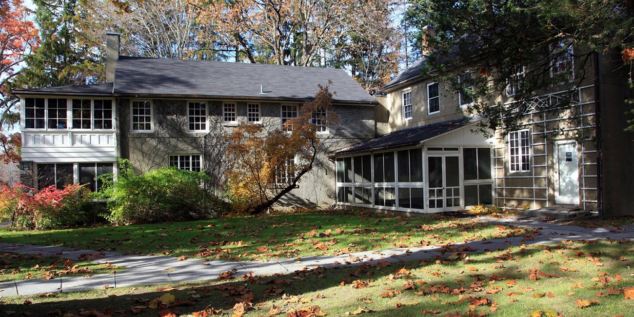 Trees and lawn surround a two-story, L-shaped stone cottage with several enclosed porches.