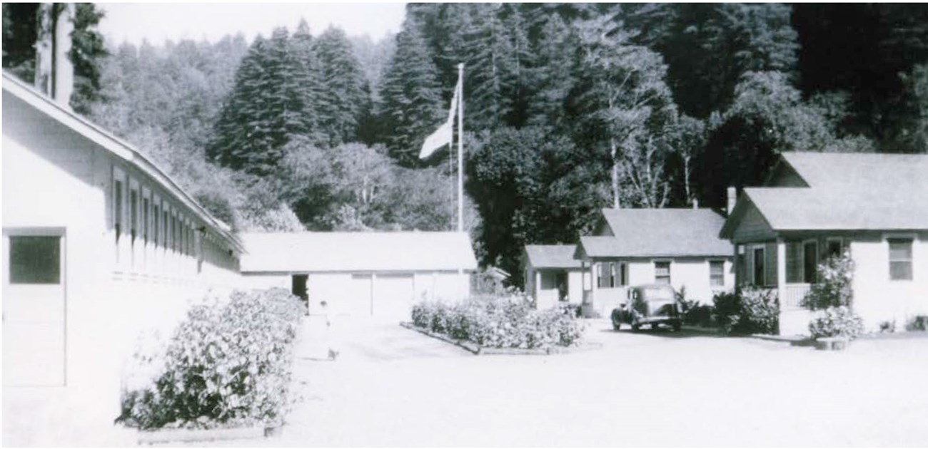 Hatchery entry view, ca. 1943 A group of one-story structures form a perimeter around a courtyard, with flower beds, a flagpole, and a car parked in front of a residence.