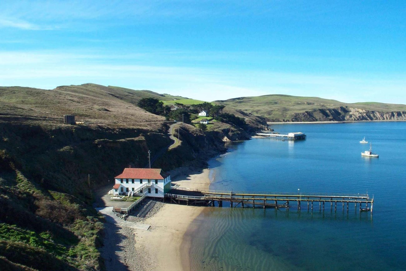 Modern View of Historic Lifeboat Station at Point Reyes National Seashore. Overhead view of a coastline, with exposed bluffs dropping to calm water. A long wharf extends into the water from a building.