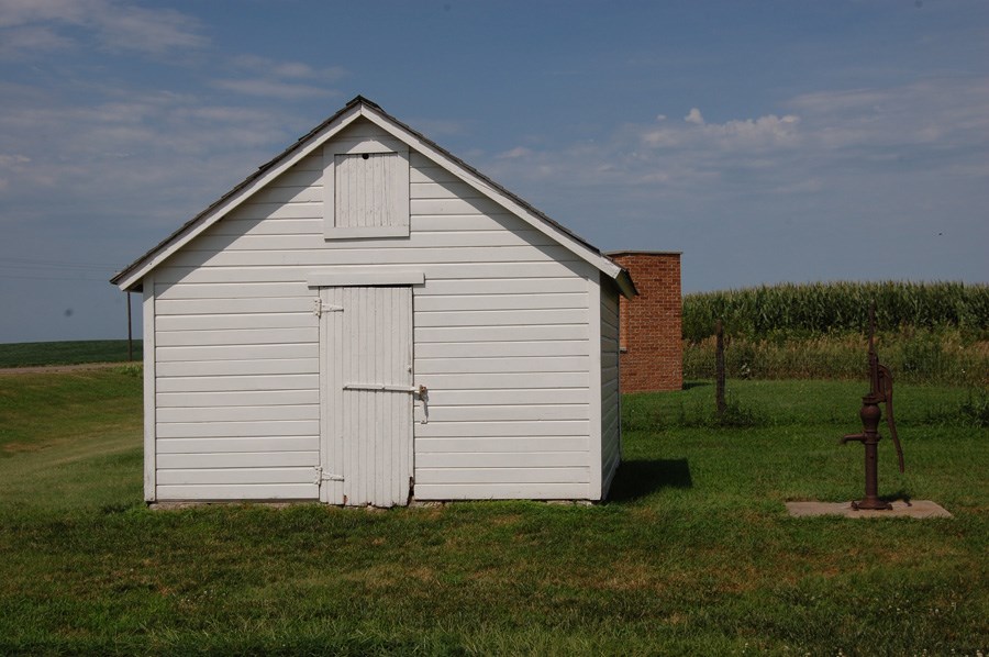 A water pump beside a wooden storage shed at Freeman School, surrounded by short grass