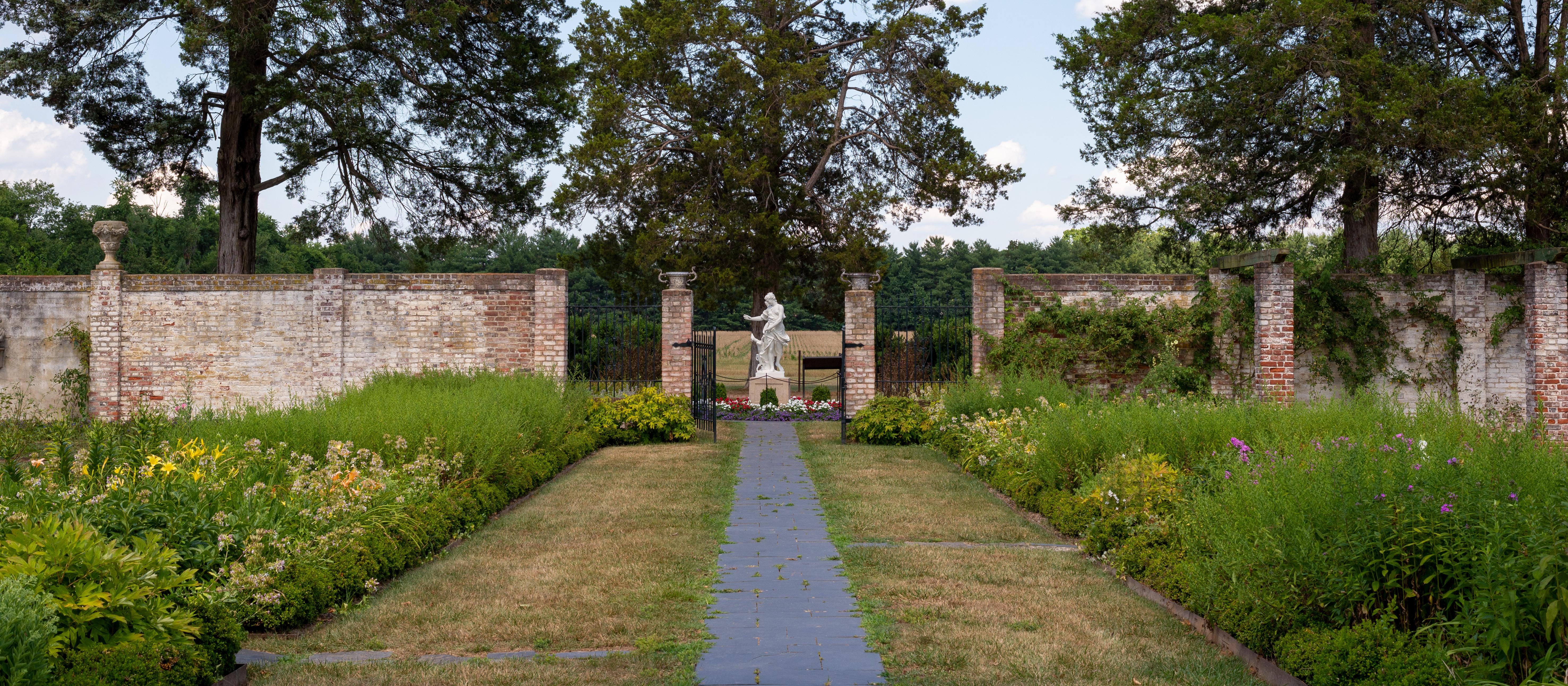 Plants and flowers grow in garden beds on both sides of a walkway, leading to a brick wall and fields beyond