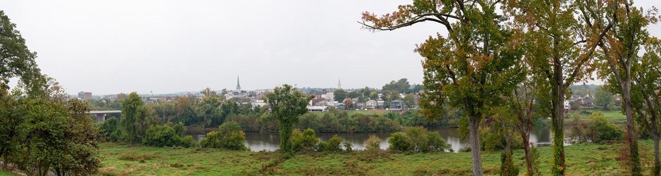 Fredericksburg from Chatham Manor View from a high point across a river bordered by leafy trees to a small town under a cloudy sky.