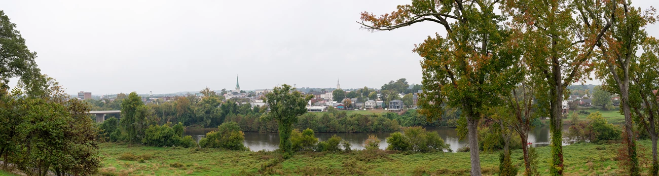 Fredericksburg from Chatham Manor View from a high point across a river bordered by leafy trees to a small town under a cloudy sky.
