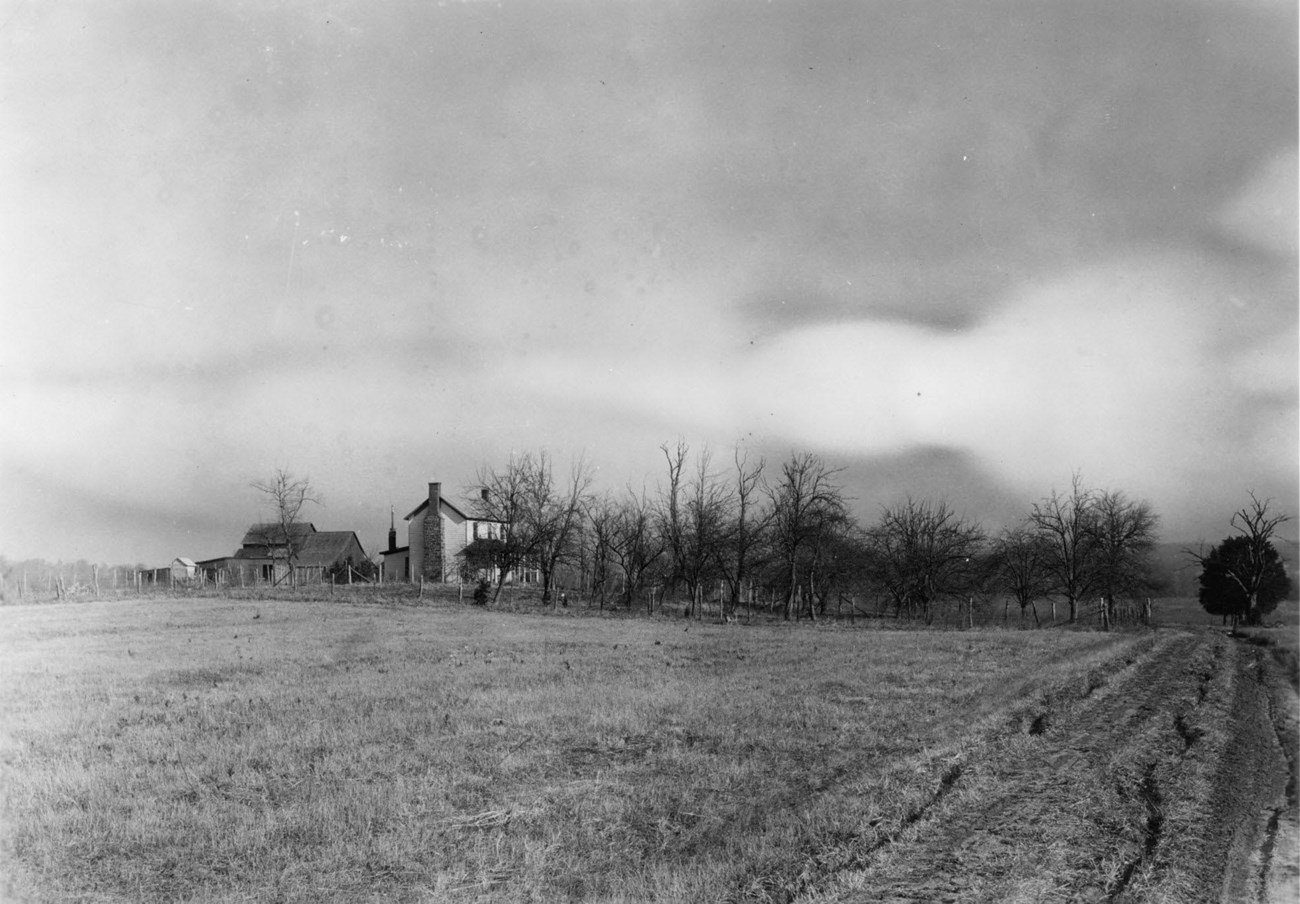 A black and white photograph showing a field in the foreground and a stand of trees and a number of farm buildings in the middle ground.